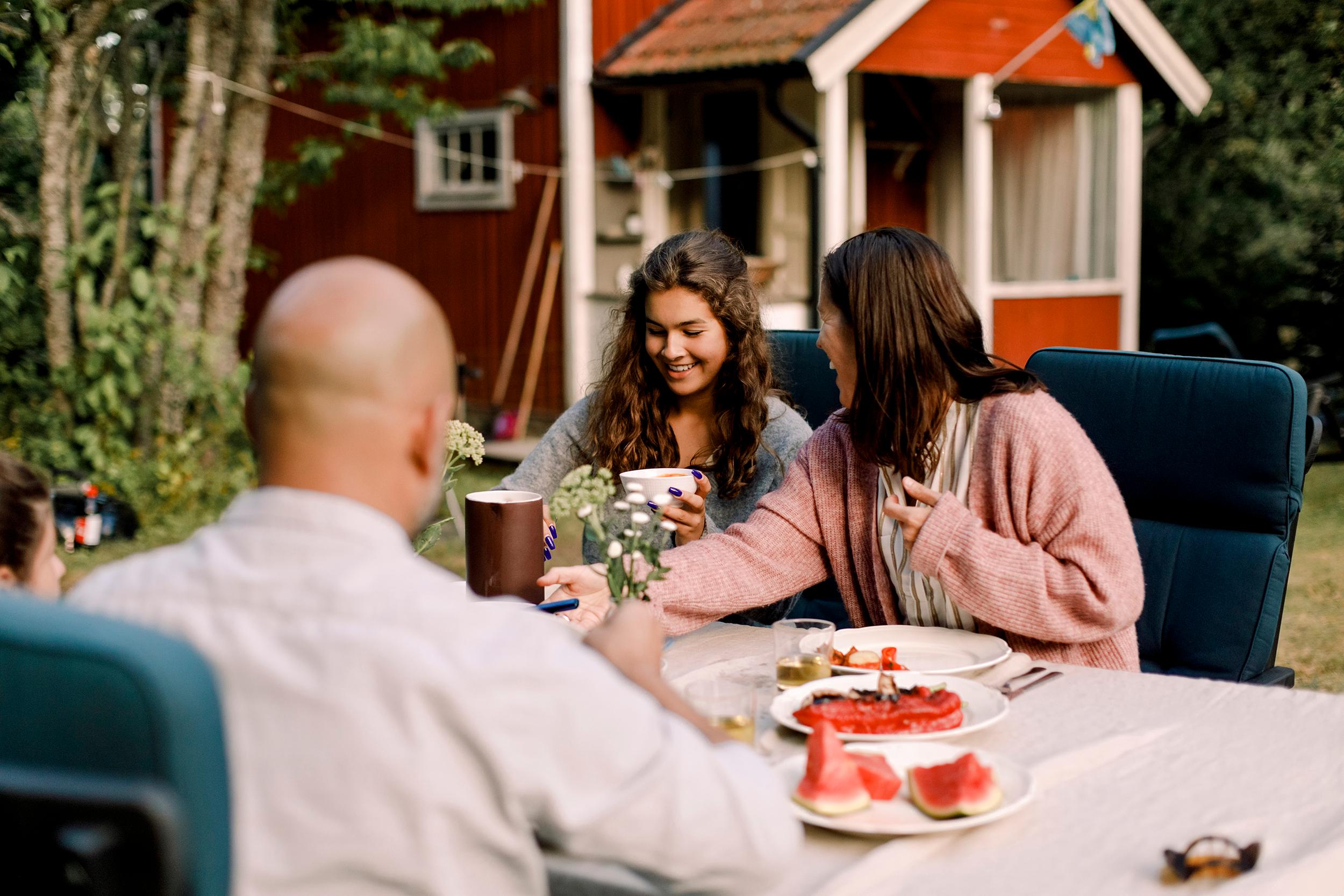 Perhe syö vesimelonia ulkona mökin edustalla pöydän ääressä. Ruskea hiuksiset äiti ja tytär hymyilevät toisilleen. Kalju isä istuu naama heitä kohden. 