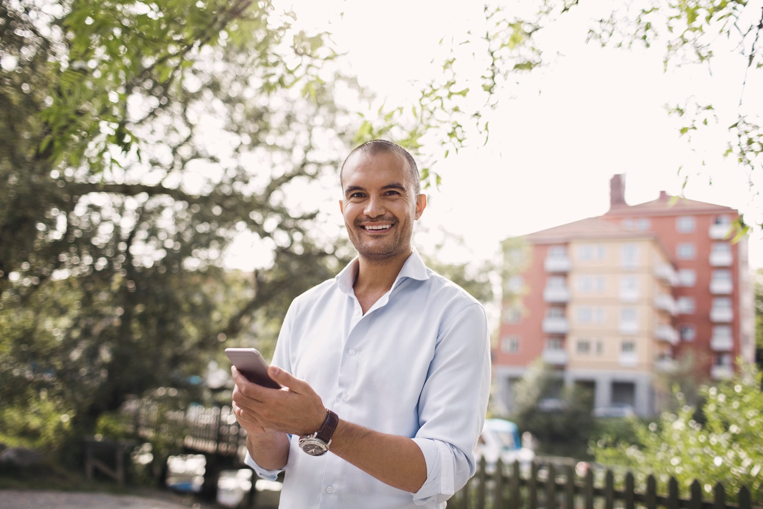 Vaaleaan kauluspaitaan pukeutunut mies pitelee älypuhelintaan ja hymyilee katse suoraan kameraan. Mies seisoo vehreässä kaupunkiympäristössä joen varrella, taustalla näkyy kelta-punainen kerrostalo. 
