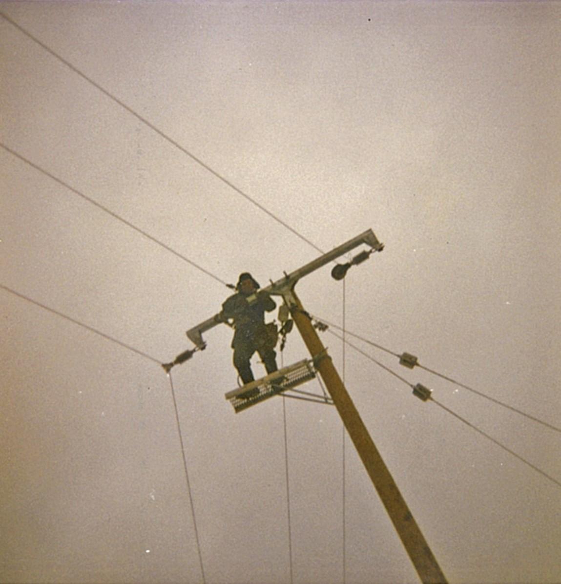 Kuvakaappaus vanhasta valokuvasta 1970-1980-luvulta. Kuvassa näkyy sähköasentaja, joka työskentelee ylhäällä pylväässä. Kuva on otettu alaviistosta. 