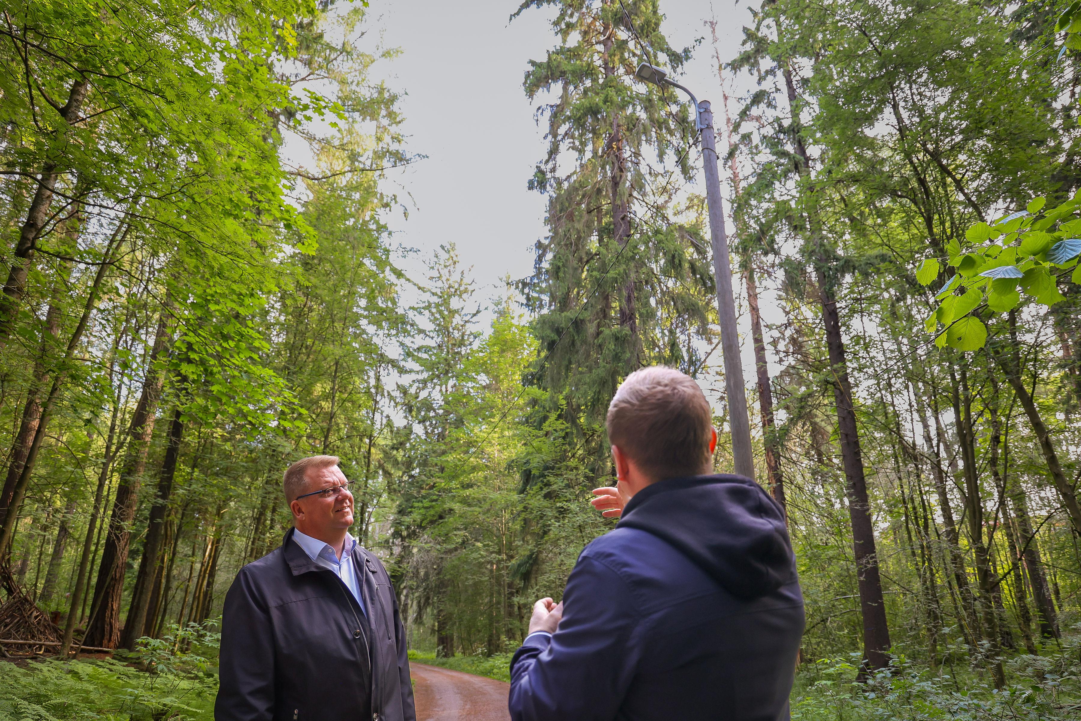 Kaksi miestä keskustelee suomalaisen metsätien varrella. Toinen miehistä seisoo selkä kameraan päin ja osoittaa taustalla näkyvää ulkovalaisinta. 