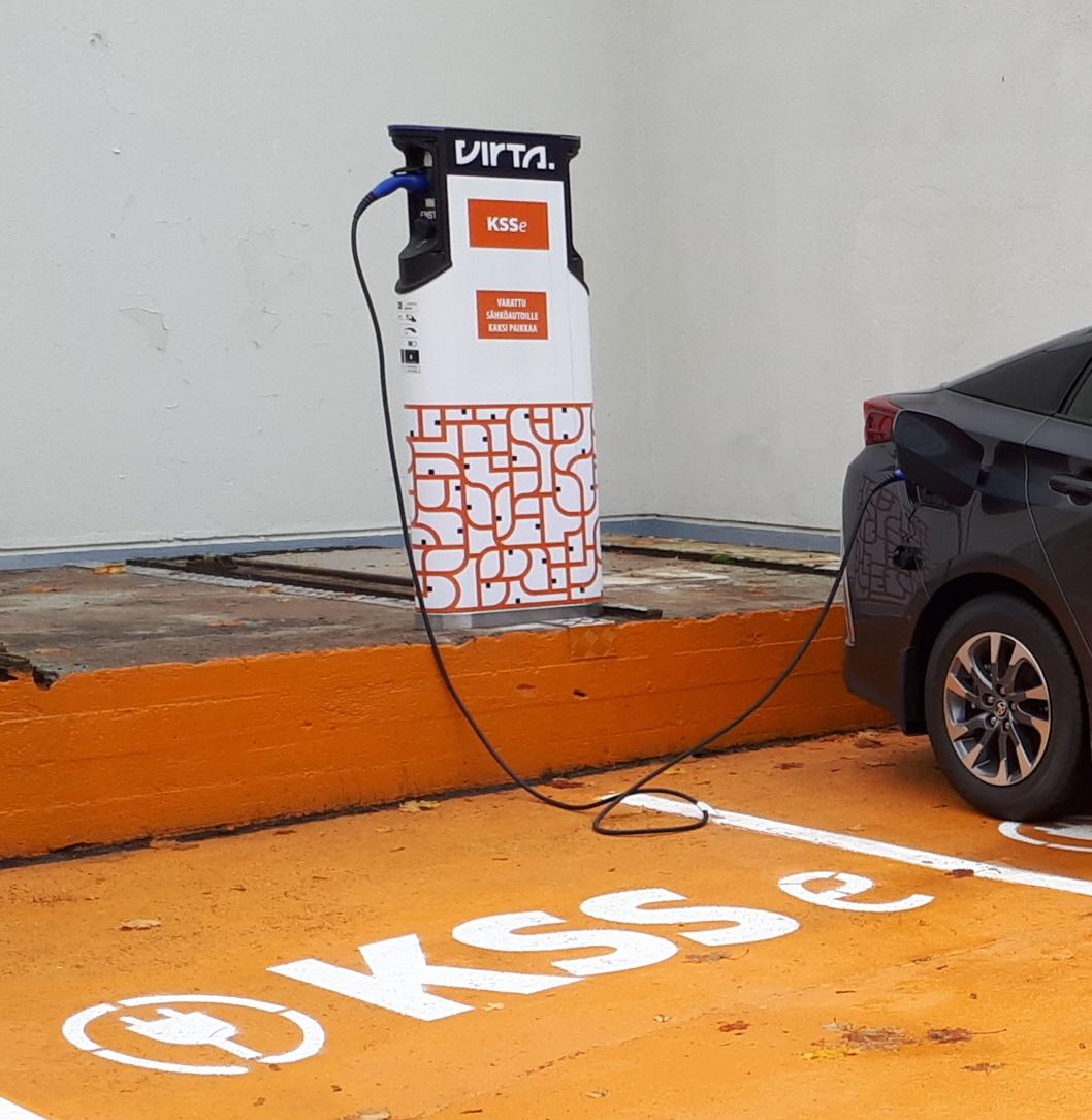 KSSe-sähköauton latauspiste, jossa on musta sähköauto latauksessa. Taustalla harmaa rakennus, parkkiruuduissa oranssi pohja ja KSSe-teksti valkoisella. 