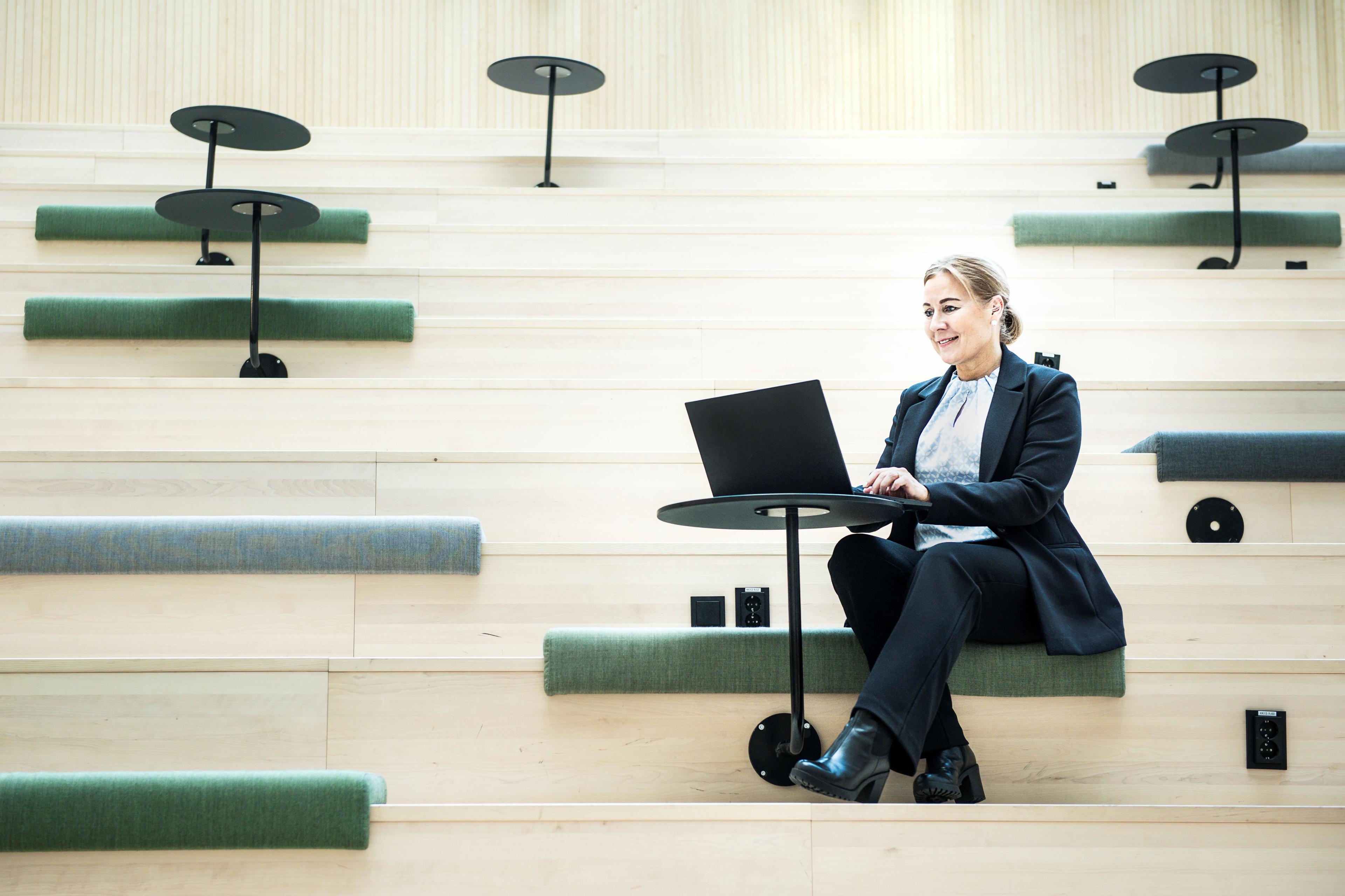 Tietokoneella työskentelevä vaaleahiuksinen nainen istuu skandinaavisen tyylisessä vaaleaa puuta olevassa portaikossa, jossa on vihreän sävyisiä istuinpehmusteita. Vaaleaan paitaan, tummiin housuihin ja jakkuun pukeutunut nainen katsoo suoraan tietokoneen näyttöön.