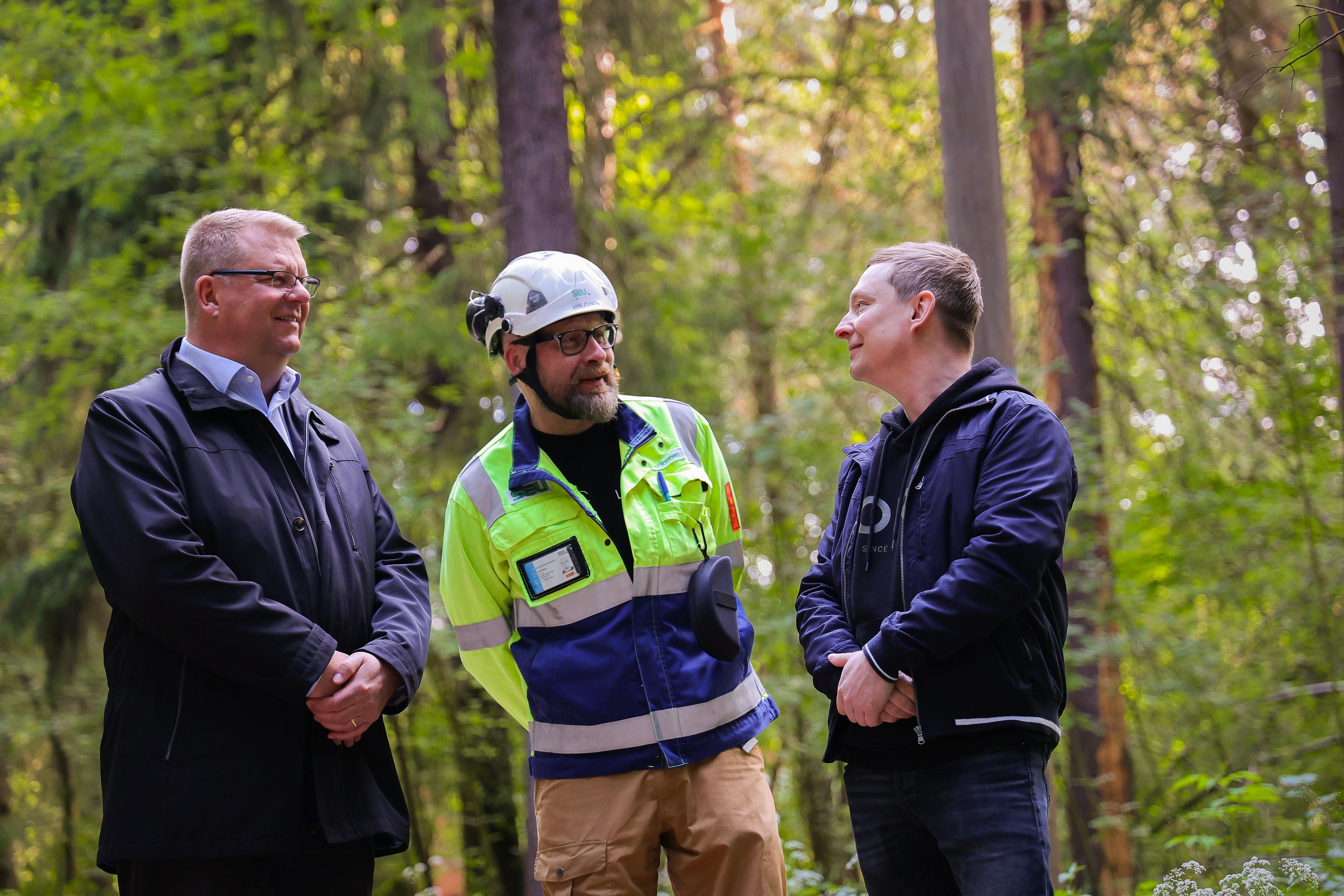 Kolme miestä keskustelee metsämaisemassa kasvot kameraa kohti. Vasemmalla olevalla miehellä on kauluspaita ja tumma takki, keskimmäisellä miehellä on valkoinen työmaakypärä ja työmaatakki, oikean puolimmaisella miehellä on sininen vapaa-ajan takki. Taustalla näkyy kesäistä suomalaista metsämaisemaa, koivuja, mäntyjä ja kuusia.