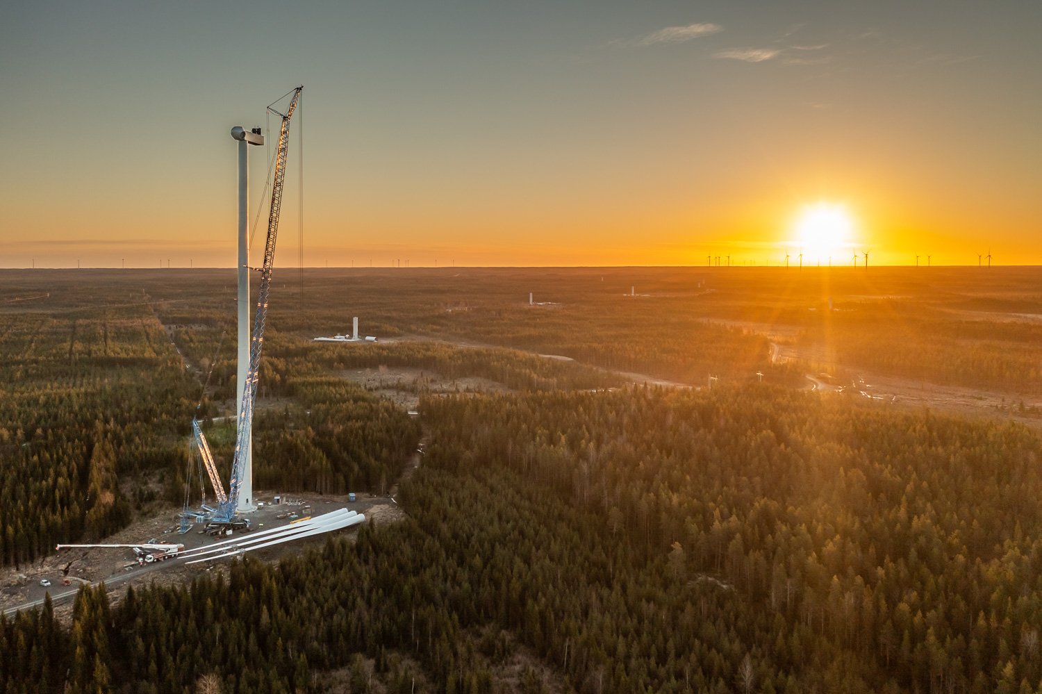 Ilmakuvassa on tuulivoimalan rakentaminen menossa, siinä näkyy tuulimyllyn runko ja nosturi. Taustalla on paljon metsämaisemaa ja laskeva aurinko.