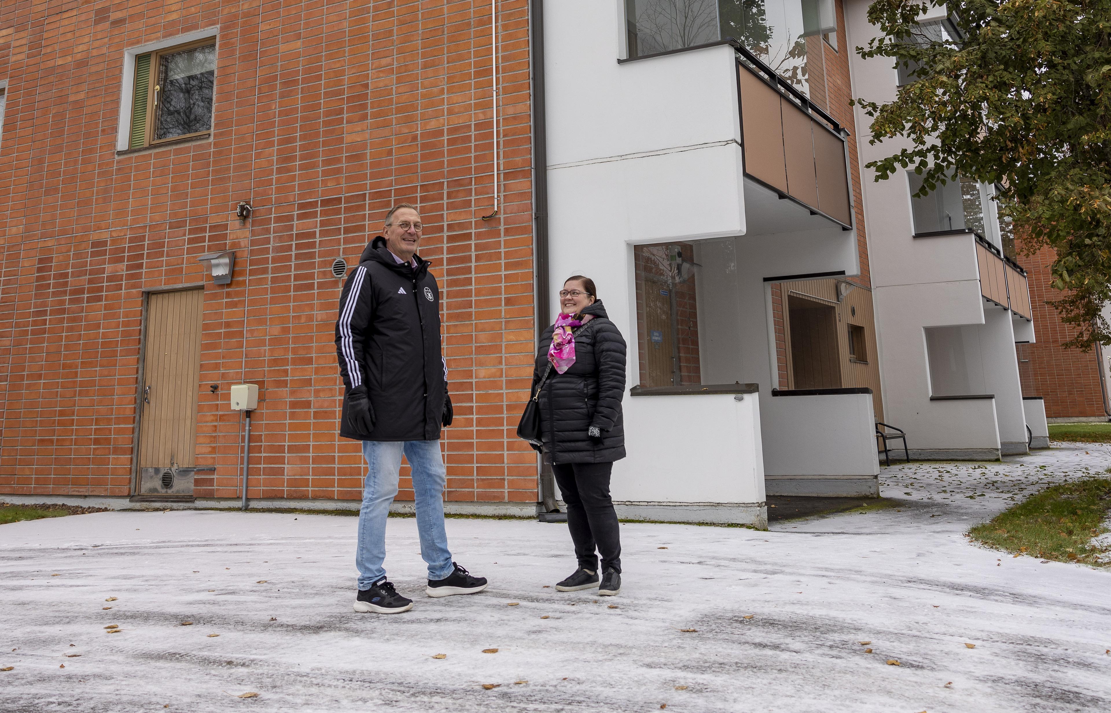 Mies ja nainen seisovat hymyilevinä punatiilisen kerrostalon edustalla alkutalven säässä. Molemmilla on päällään mustat toppatakit, maassa on pieni kerros lunta. Taustalla näkyvässä kerrostalossa on valkoiset parvekkeet. Pihassa on suuri vaahtera, joka näkyy kuvan toisella laidalla.