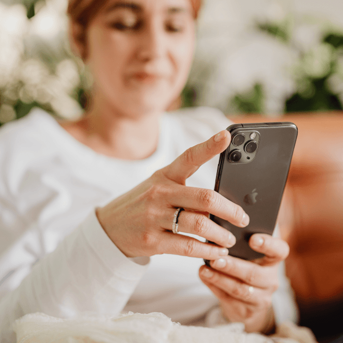 Keski-ikäinen nainen tutkii harmaata Apple-merkkistä puhelinta. Kuva kohdistettu käsissä olevaan puhelimeen. Naisella lyhyet punaruskeat hiukset ja vaalea pitkähihainen neulepusero. Taustalla näkyy ruskea nahkasohva ja viherkasveja.