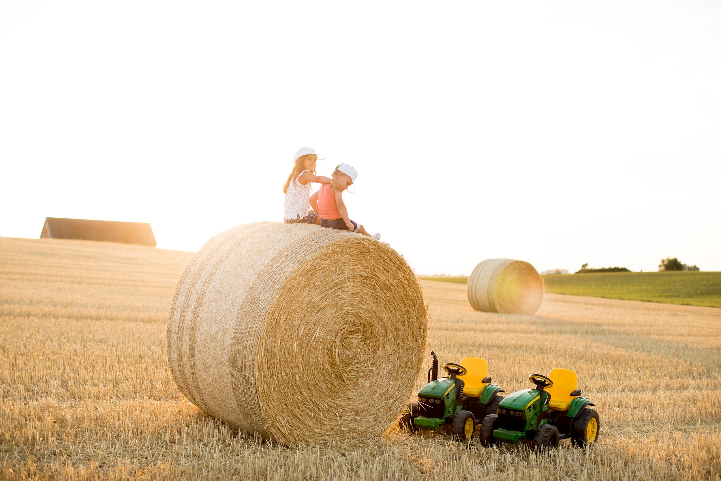 Kaksi lasta istuu keltaisella sänkipellolla suuren pyöreän heinäpaalin päällä. Pelolla on myös toinen samanlainen heinäpaali kauempana, sekä kaksi lasten vihreää ja traktoria, joissa on keltaiset penkit. Taustalla näkyy peltoa, aurinkoista vaaleaa taivasta ja ladon katto.