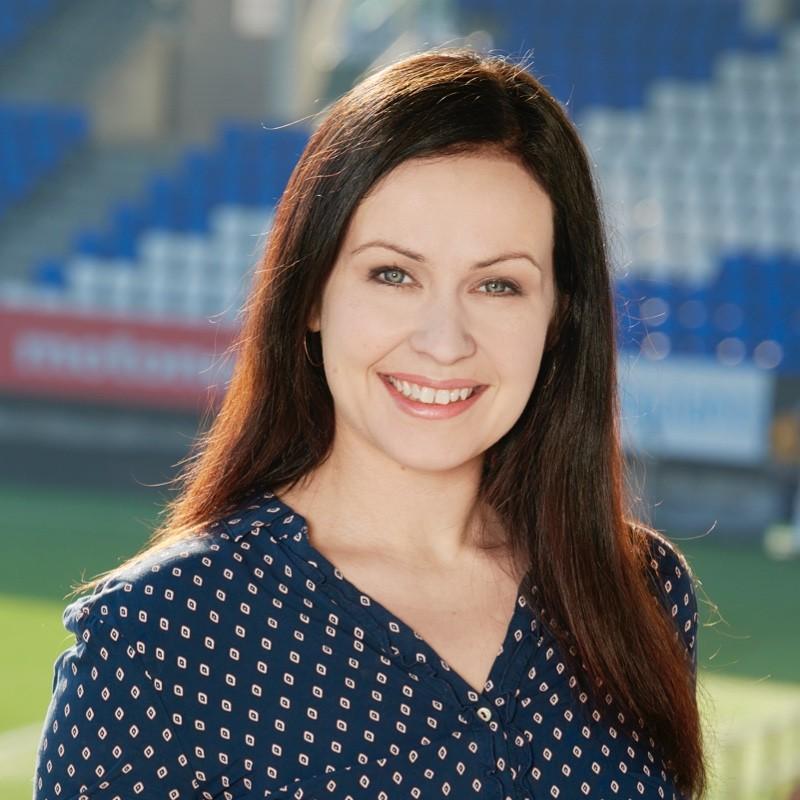 Nainen katsoo kohti kameraa. Hänellä on tummat hiukset ja siniset silmät. Päällä on pilkullinen sininen pusero. Aurinko paistaa ja taustalla näkyy jalkapallo stadionin penkkejä.