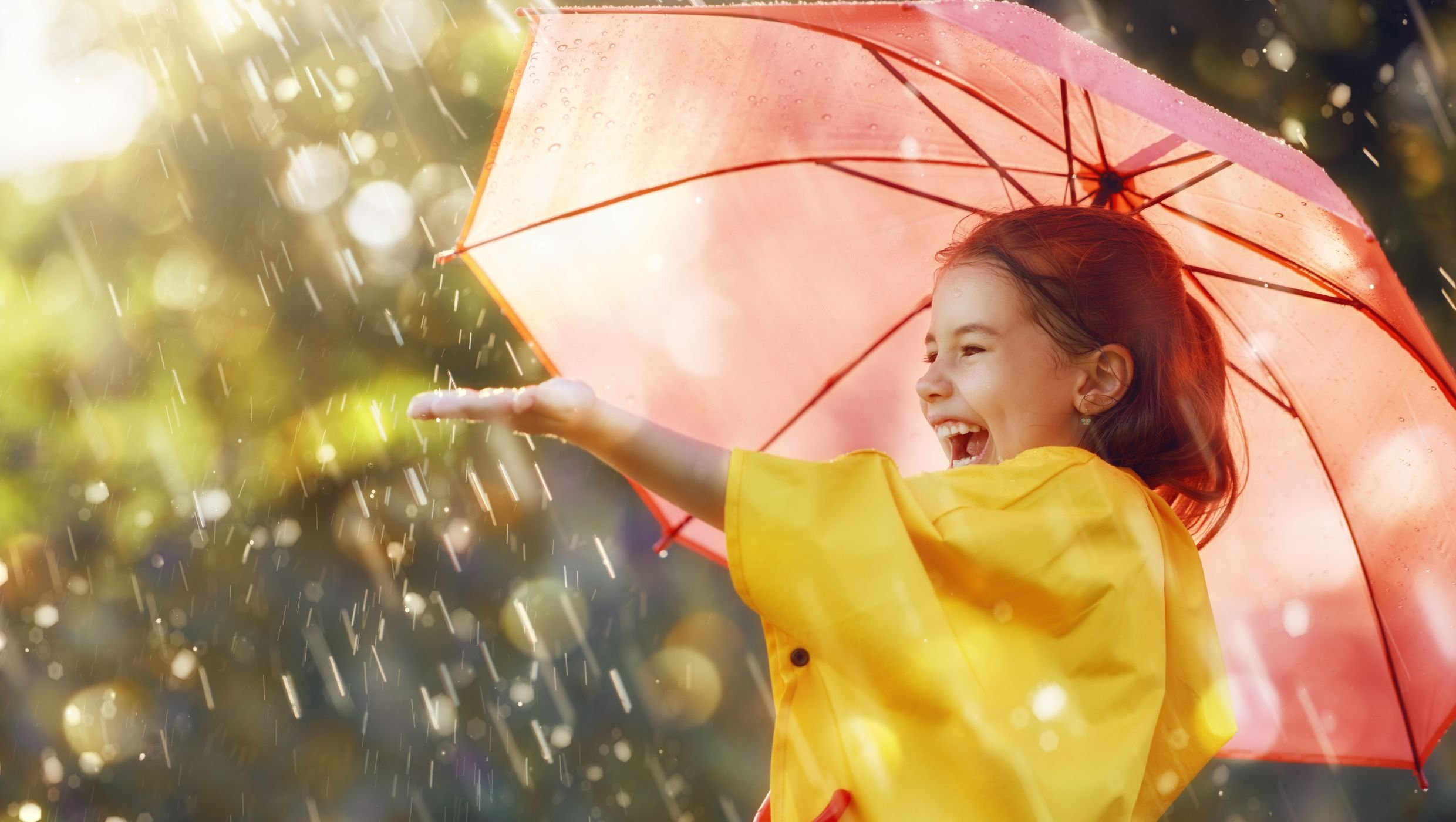 Pieni ruskeahiuksinen tyttö keltaisessa sadetakissa ja vaaleanpunaisen sateenvarjon alla ojentaa vasemman käden kohti sadepisaroita. Taustalla näkyy vihreää ja auringon pilkahduksia.