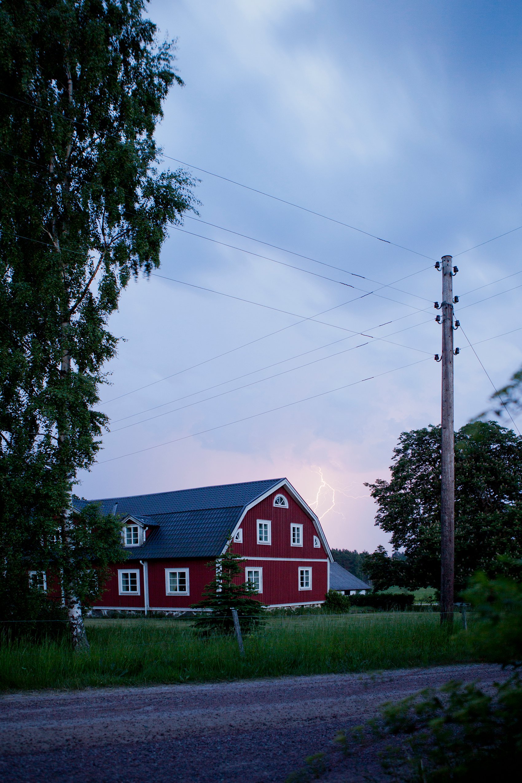 Ilmassa kulkee sähköjohtoja moneen suuntaan punaisen maalaistalon lähistöllä, myrskyisellä taivaalla näkyy salamanisku.