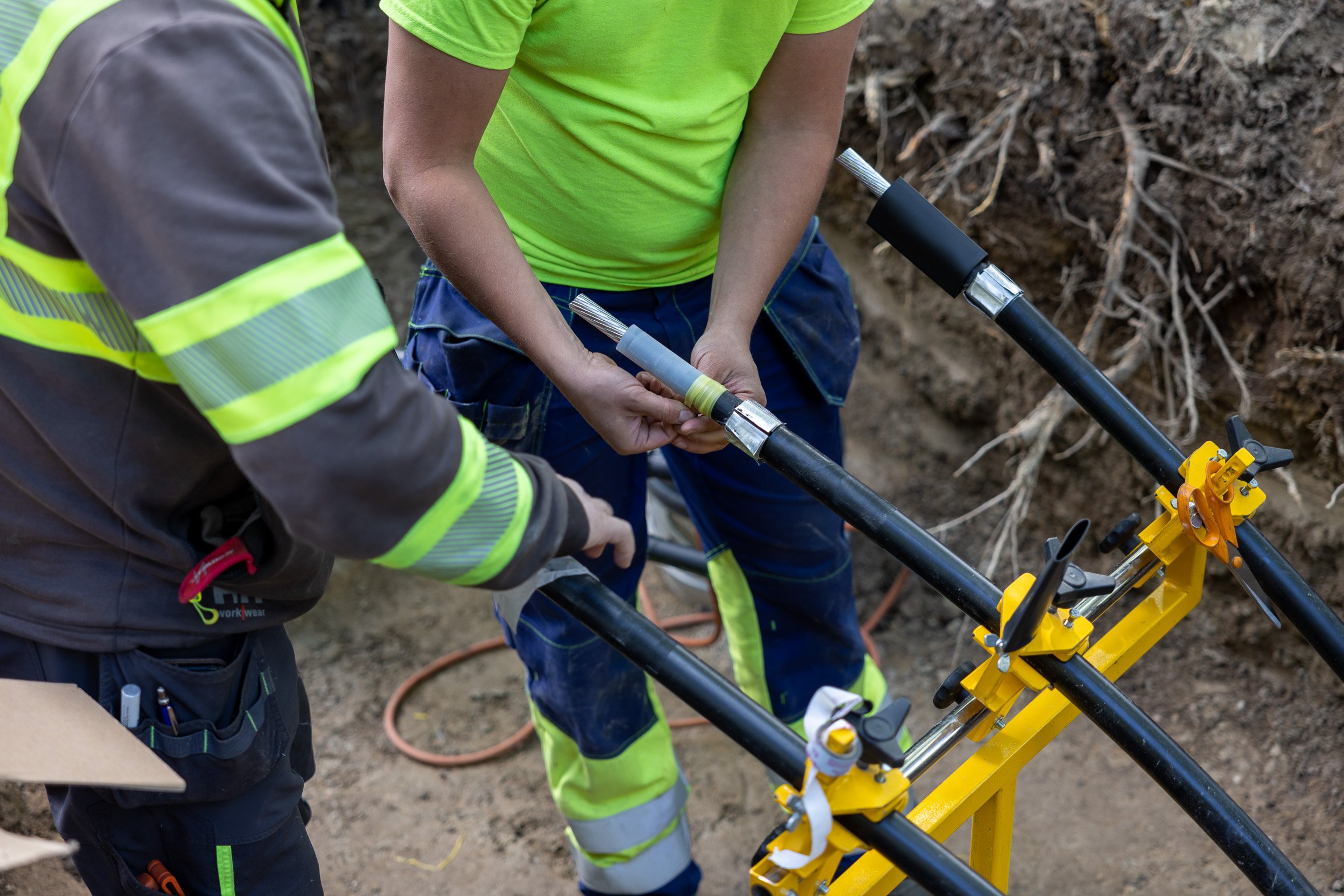 Syvän kaivannon pohjalla olevaan keltaiseen metallitelineeseen on kiinnitetty rinnakkain kolme mustaa kaapelia, joita kaksi työasuista henkilöä käsittelevät.