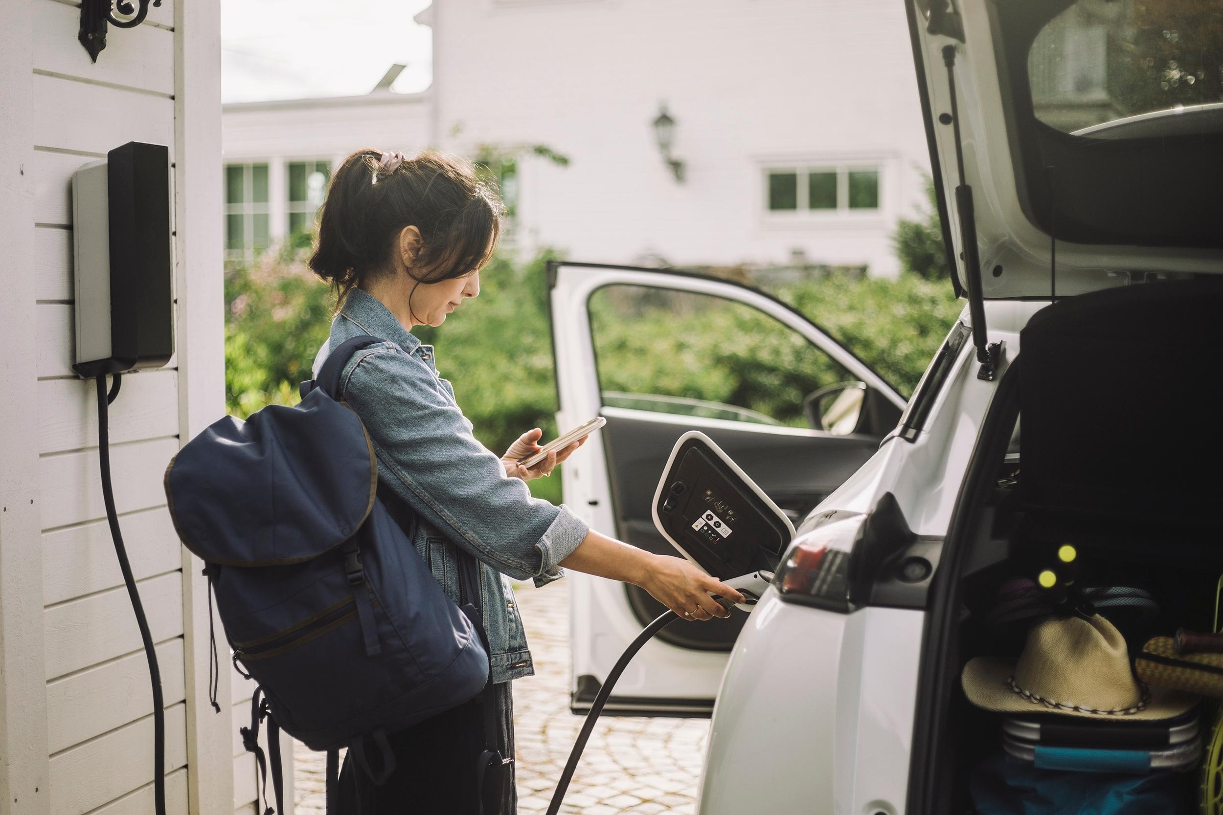 Nainen tutkii kädessään olevaa puhelinta ja laittaa sähköauton latauspistoketta autoon kiinni.