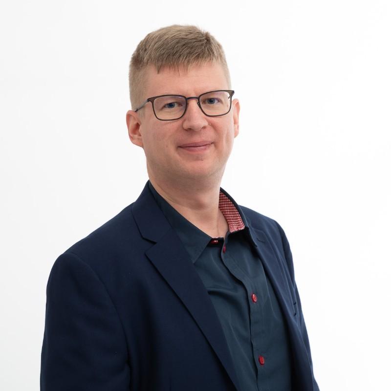 Potrettikuva miehestä. Miehellä on musta puvuntakki ja kauluspaita. Hänellä on lyhyet hiukset ja mustat kulmikkaat silmälasit.
